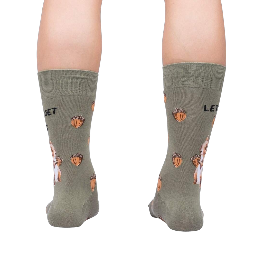 A pair of crew socks worn on legs, shown from the back. The socks are olive green with a pattern of orange acorns, white rabbits, and black text reading 'LET GET'.