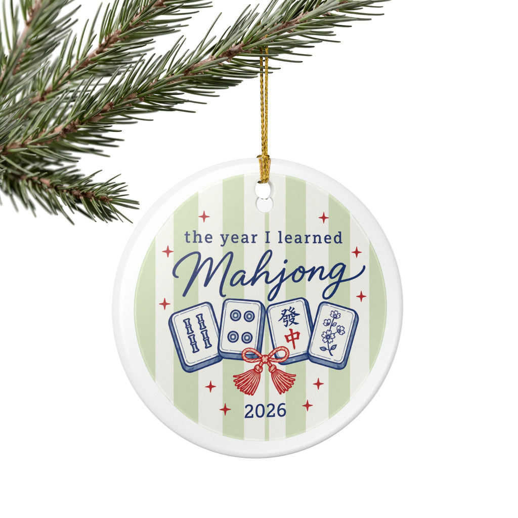 A round white ceramic Christmas ornament hanging from a pine branch. The ornament features a light green and white striped background with the text 'the year I learned Mahjong 2026' and illustrations of four mahjong tiles tied with a red bow.