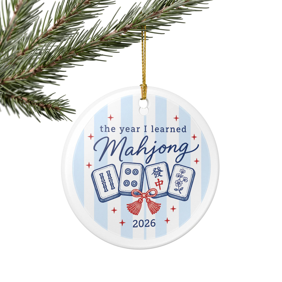 A round white ceramic Christmas ornament hanging from a gold string on a pine branch. The ornament features a light blue and white striped background with the text 'the year I learned Mahjong 2026' and illustrations of four mahjong tiles tied with a red bow.