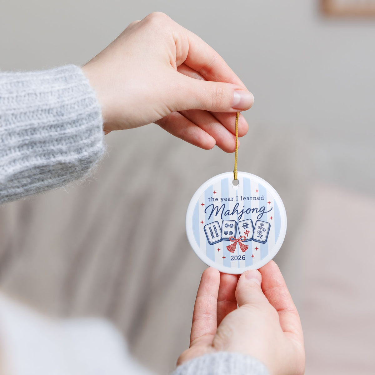A person holding a round white Mahjong-themed ornament with a gold hanging string. The ornament features blue text reading 'the year I learned Mahjong 2026' and illustrations of four playing cards on a light blue striped background.