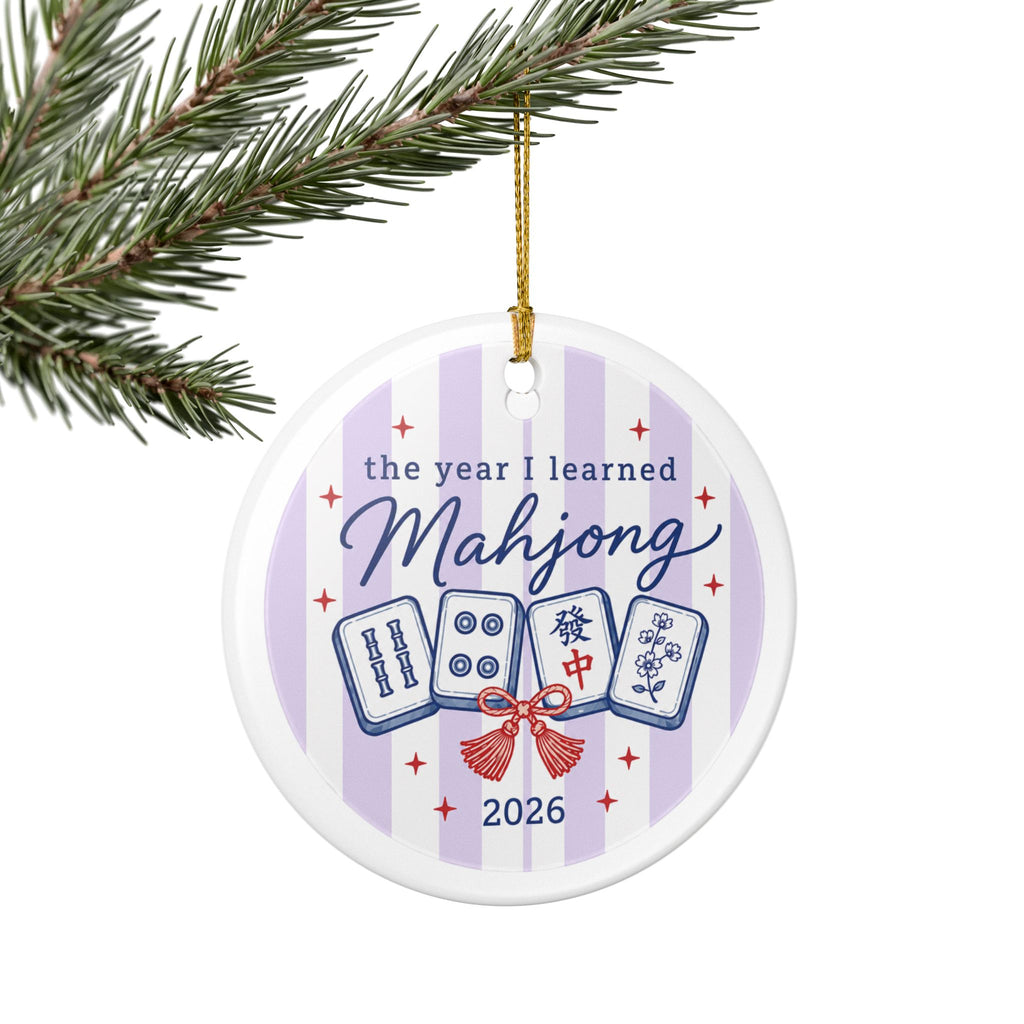 A round white ceramic Christmas ornament hanging from a gold string on a pine branch. The ornament features a light purple and white striped background with the text 'the year I learned Mahjong 2026' and illustrations of four mahjong tiles tied with a red bow.
