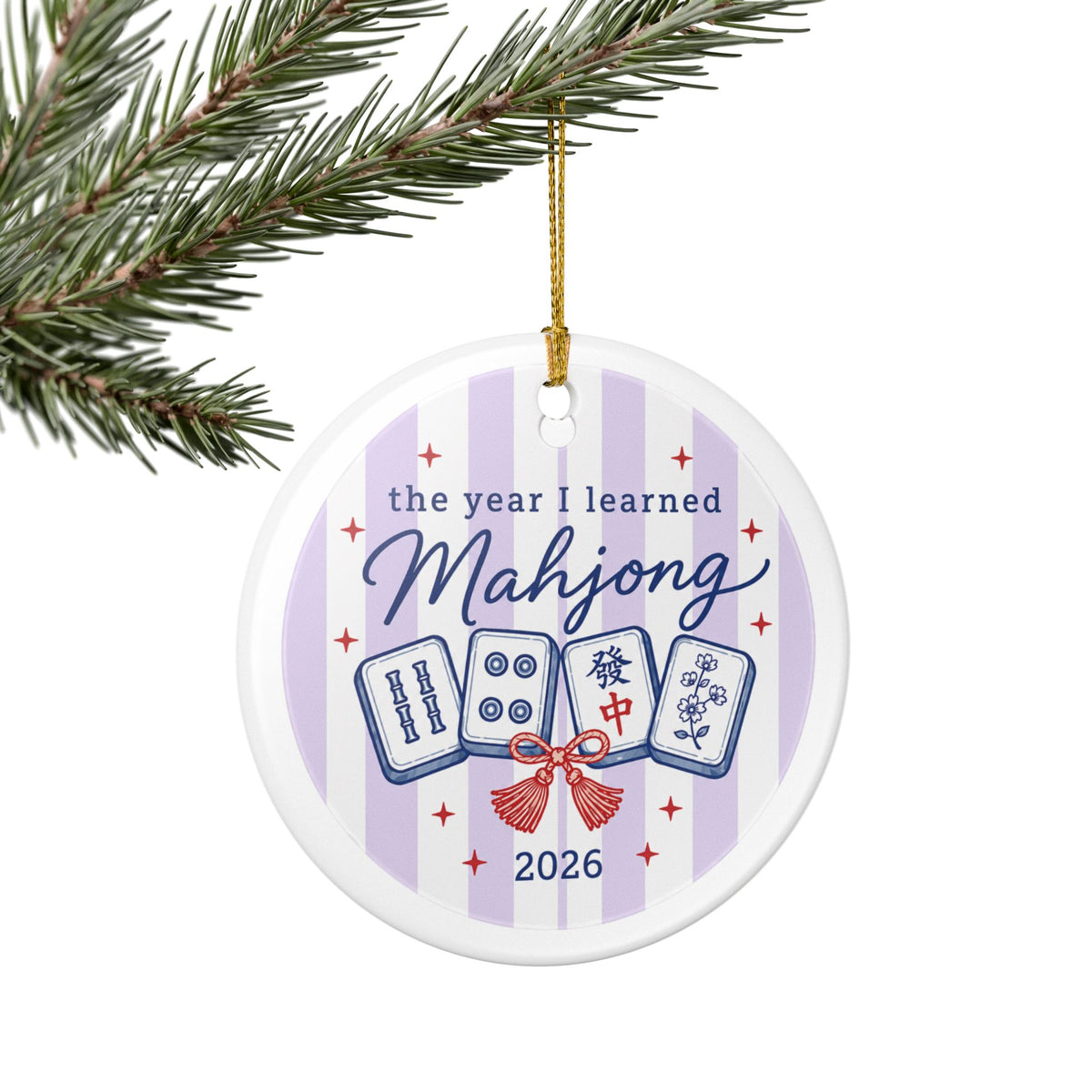 A round white ceramic Christmas ornament hanging from a gold string on a pine branch. The ornament features a light purple and white striped background with the text 'the year I learned Mahjong 2026' and illustrations of four mahjong tiles tied with a red bow.