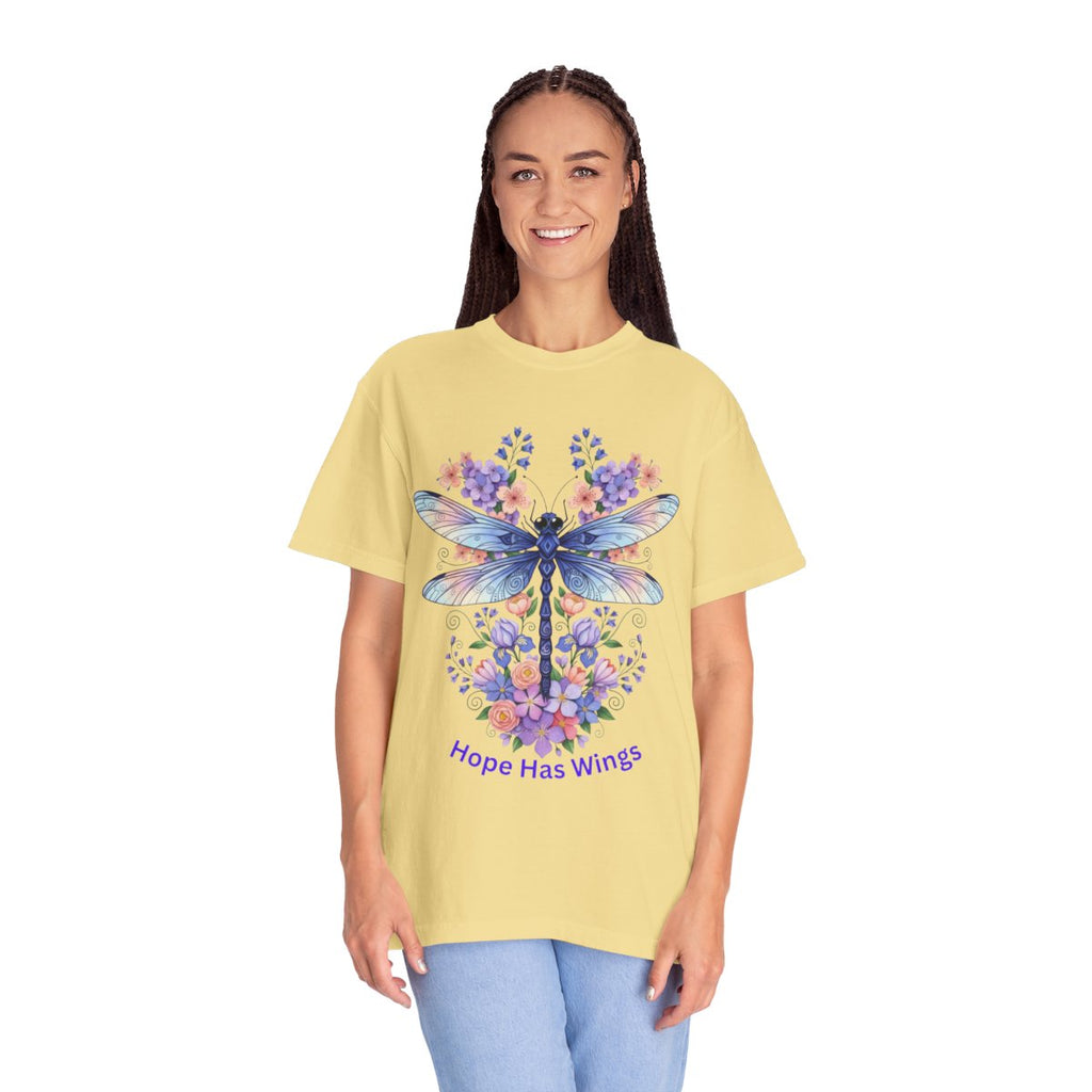 A woman modeling a pale yellow graphic t-shirt featuring a blue dragonfly surrounded by purple and blue flowers, with the text 'Hope Has Wings' printed below. She is wearing light blue jeans against a solid white background.