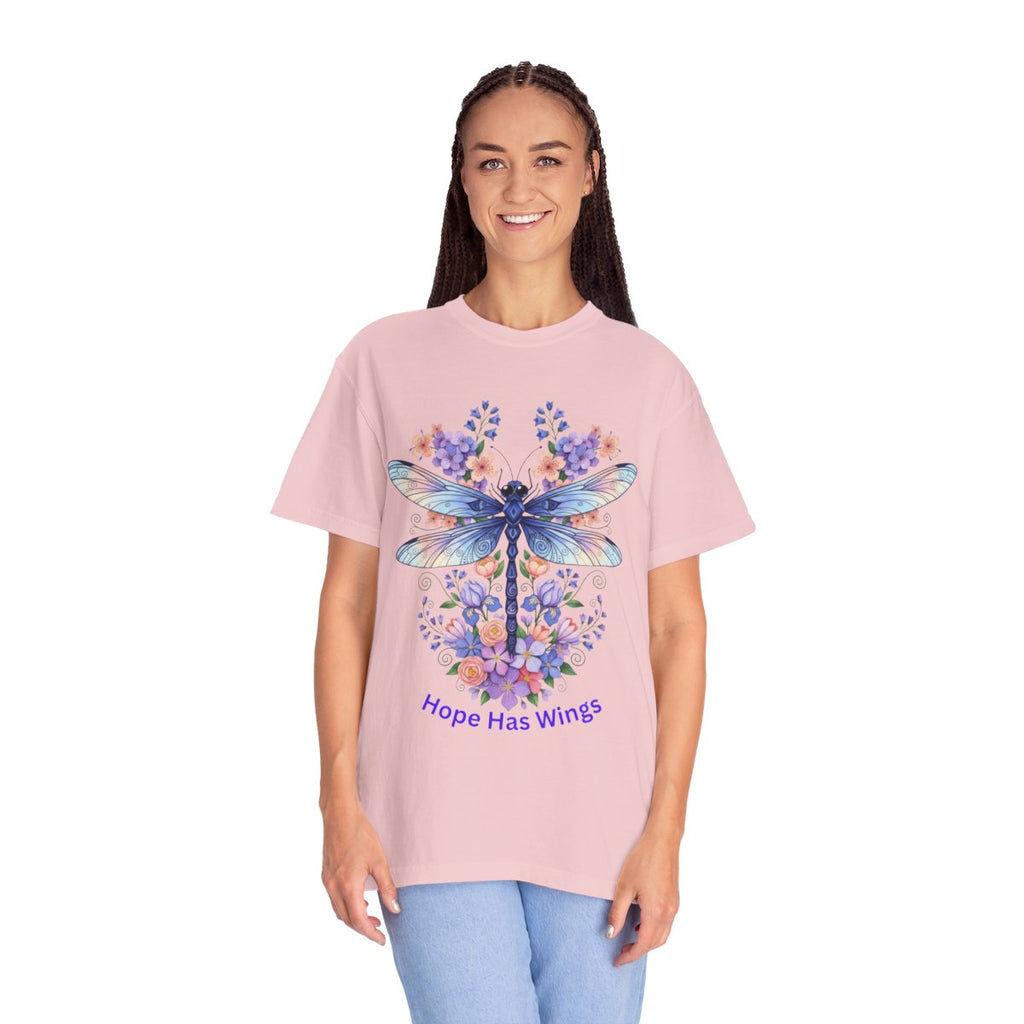 A woman modeling a light pink short-sleeve t-shirt with a large graphic of a blue dragonfly surrounded by purple and blue flowers, with the text 'Hope Has Wings' printed below. She is wearing light blue jeans against a plain white background.