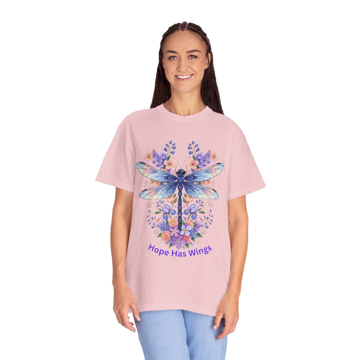 A woman modeling a light pink short-sleeve t-shirt with a large graphic of a blue dragonfly surrounded by purple and blue flowers, with the text 'Hope Has Wings' printed below. She is wearing light blue jeans against a plain white background.